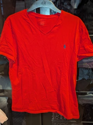 Röd v-ringad t-shirt från Polo Ralph Lauren - Röd t-shirt från Polo Ralph Lauren med v-ringning och den klassiska blå broderade loggan på bröstet. Tillverkad i mjuk bomull och har korta ärmar. Inga synliga fläckar eller defekter. Skapa en bundle för bättre pris