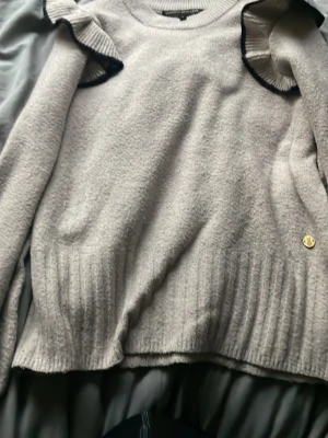 Beige stickad tröja med volang - Supersöt beige stickad tröja från Pernilla Wahlgren med volangdetaljer vid axlarna och svarta kanter. Tröjan har rund halsringning, långa ärmar och ribbstickade muddar. Perfekt för dig som gillar en feminin och trendig stil.