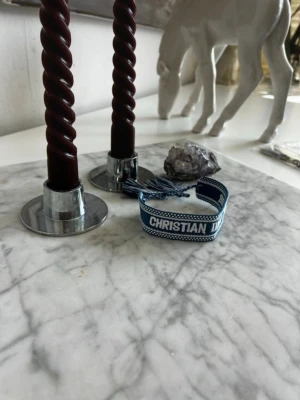 Blått Christian Dior tygarmband - Snyggt blått tygarmband från Christian Dior med texten 'J'ADIOR' broderat i vitt. Armbandet har justerbar knytning med tofsar i ändarna och en cool, ungdomlig design. Perfekt accessoar för att lyfta din outfit med en exklusiv touch.
