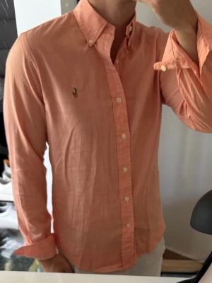 Persikofärgad skjorta från Ralph Lauren - Klassisk persikofärgad skjorta från Ralph Lauren med button-down krage och broderad logga på bröstet. Skjortan har långa ärmar, normal passform och är tillverkad i bomull. Perfekt för en clean och stilren look.