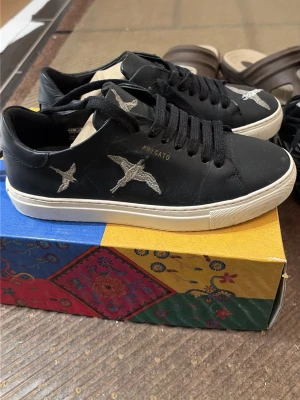 Arigato sneakers äkta  - Snygga svarta sneakers från Axel Arigato med broderade fåglar i silver på sidan och tån. Skorna har svart snörning, vit sula och är tillverkade i slätt läder. Perfekta för dig som vill ha en clean men ändå unik look.