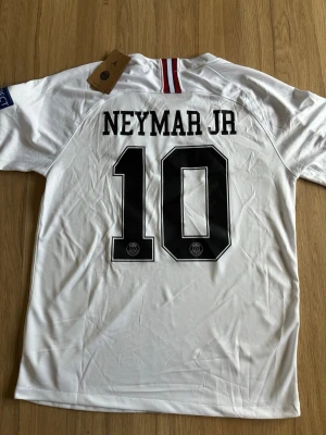 Vit PSG Neymar JR 10 matchtröja - Säljer en vit PSG t-shirt med Neymar JR och nummer 10 på ryggen. Framsidan har Fly Emirates-tryck, PSG-logga och Jordan-logga. Kortärmad modell i lätt och skönt material, perfekt för fotboll eller streetstyle.