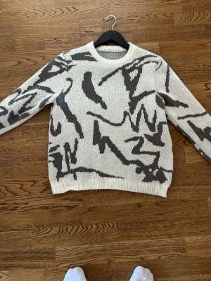 Vit stickad tröja med svart mönster - Säljer en vit stickad tröja med abstrakt svart mönster. Tröjan har rund halsringning, långa ärmar och ribbade muddar vid ärmslut och nederkant. Perfekt för dig som gillar en unik och trendig look. 158