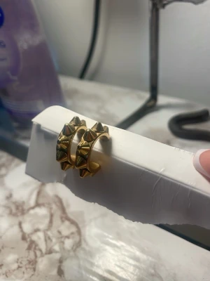 Guldpläterade örhängen med nitar - Säljer ett par chunky örhängen i guldpläterad metall med coola pyramidformade nitar runt hela ringen. De har en modern och edgy look som verkligen sticker ut. Perfekta för dig som vill addera lite attityd till din stil.