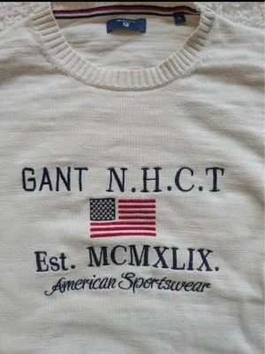 Vit tröja från Gant med USA-flagga - Vit långärmad tröja från Gant med broderad amerikansk flagga och texten 'N.H.C.T', 'Est. MCMXLIX.' samt 'American Sportswear' på bröstet. Tröjan har rund halsringning och ribbade muddar. Perfekt för en sportig och avslappnad stil.