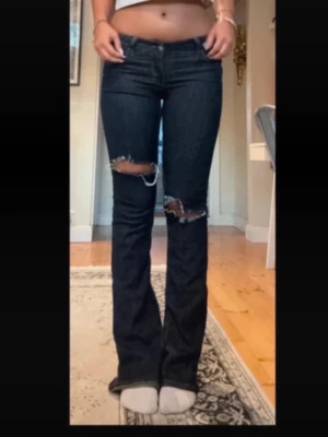 Mörkblå bootcut jeans med slitningar - Säljer ett par mörkblå bootcut jeans med slitna detaljer över båda knäna. Jeansen har klassisk femficksdesign och är tillverkade i stretchigt denimtyg som sitter snyggt på kroppen. Perfekta för en trendig och avslappnad look.