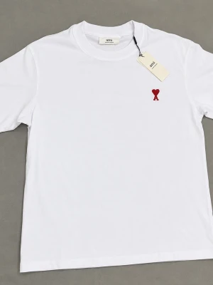 Vit t-shirt från Ami med rött hjärta - Klassisk vit t-shirt från Ami med ett litet rött hjärta broderat på bröstet. T-shirten har rund hals och korta ärmar, tillverkad i mjuk bomull för en skön känsla. Perfekt basplagg med stilren design.
