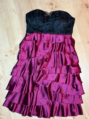 Axelbandslös vinröd volangklänning - Snygg axelbandslös klänning med svart spetsliv och volangkjol i vinrött satinliknande material. Dekorerad med en liten rosett framtill. Perfekt för dig som vill sticka ut med en elegant look.           🤎 BUDAS UT PÅ LIVE! 🤎 Denna kommer att budas ut på min Plick Live 25 augusti kl. 19:00 🛍️