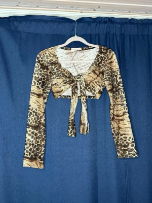 Leopardmönstrad croppad topp med knyt - Säljer en croppad långärmad topp med leopardmönster i beige och bruna toner. Toppen har v-ringning och knyt framtill som ger en snygg detalj. Perfekt för dig som vill sticka ut med ett djurmönster och trendig passform.                         🤎 BUDAS UT PÅ LIVE! 🤎 Denna kommer att budas ut på min Plick Live 25 augusti kl. 19:00 🛍️