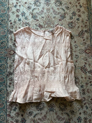 Ljusrosa ärmlös blus med rynk - Söt ljusrosa blus i ett tunt, mjukt tyg med rynkdetaljer över bröstet och midjan. Blusen är ärmlös och har en rund halsringning. Perfekt att styla med jeans eller kjol för en fräsch look.