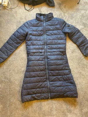 Marinblå lång dunjacka från CRW - Snygg marinblå lång dunjacka från CRW med hög krage och dragkedja hela vägen. Jackan har quiltad design, två sidofickor med dragkedja och är perfekt för kalla vinterdagar. Materialet är lätt och värmande, och modellen är figurnära. perfekt att ha när man rider.