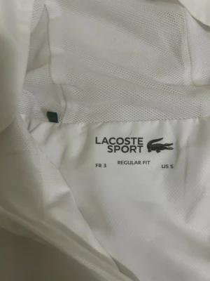 Vit vindjacka från Lacoste Sport - Snygg vit vindjacka från Lacoste Sport med regular fit. Jackan har dragkedja framtill, meshfoder och den klassiska gröna krokodilloggan på bröstet. Perfekt för dig som vill ha en stilren och sportig look. Materialet är lätt och andas bra och användes ett fåtal gånger 