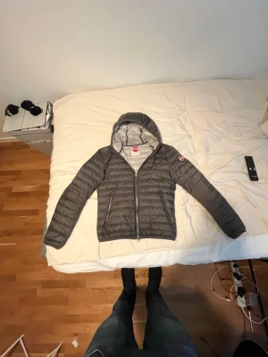Grå dunjacka med huva från Colmar - Snygg grå dunjacka från Colmar med huva och dragkedja framtill. Jackan har quiltad design och en diskret logga på ärmen. Perfekt för kalla vinterdagar och riktigt lätt att bära. Passar dig som vill ha en stilren och funktionell jacka.