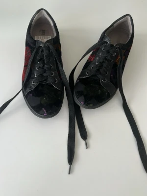 Svarta sneakers med färgglada detaljer - Unika svarta sneakers med färgglada broderade detaljer i rött, lila och orange. Skorna har svarta snören, svart sula med stjärnmönster undertill och en mix av textil och glansigt material på ovandelen. Perfekta för dig som vill sticka ut med din stil.