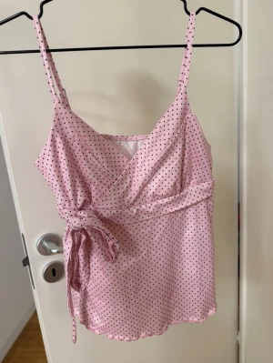 Rosa prickig pyjamastopp med knyt - Supersöt rosa topp med svarta prickar, smala axelband och omlottdesign framtill. Toppen har en knytning i sidan och volangkant både fram och bak för en extra gullig look. 