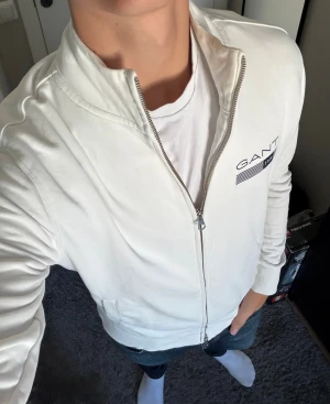Vit ziptröja från GANT - Snygg vit ziptröja från GANT med hög krage och dragkedja framtill. Tröjan har ribbade muddar och fickor på sidorna. GANT-logga med flaggdetalj på bröstet ger en clean och stilren look.