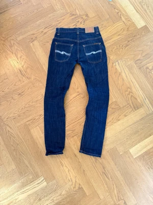 Mörkblå jeans från Nudie - Säljer ett par mörkblå jeans med smal passform och kontrastsömmar. Jeansen har två bakfickor med vit broderad detalj och klassisk femficksdesign. Materialet är denim och de har en snygg, tidlös look. Storlek 29/32!