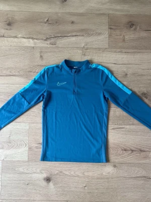 Blå långärmad träningströja Nike - Snygg blå långärmad träningströja från Nike med halv dragkedja framtill och ljusblåa detaljer längs axlarna. Perfekt för träning eller uppvärmning. Tillverkad i ett stretchigt funktionsmaterial som andas.