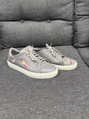 Axel Arigato  - Snygga grå sneakers i läder med färgglada broderade fåglar på sidan och framtill. Skorna har vita sulor och klassisk snörning. Perfekta för dig som vill ha en unik detalj på dina skor.