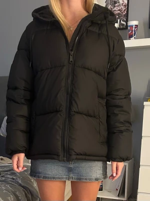 Svart vinterjacka med huva - Säljer en svart pufferjacka med huva och dragkedja framtill. Jackan har en quiltad look och är tillverkad i syntetmaterial, perfekt för kalla dagar. Den har lång ärm och en oversized passform som ger en chill vibe.