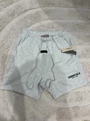  Essentials shorts - Snygga ljusgrå shorts från Essentials med elastisk midja och snörning framtill. Mjuk bomullskänsla och diskret logga på vänster ben. Perfekta för chill dagar eller till träningen.