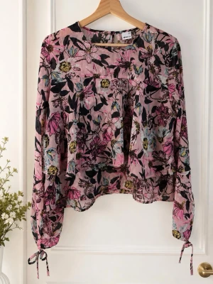 Blommig rosa blus från Gina Tricot - Säljer en luftig blommig blus från Gina Tricot i storlek 44. Blusen är rosa med färgglada blommor i svart, gult och blått, har långa ärmar med knytband vid handlederna och är gjord i ett tunt, transparent material. Perfekt för en trendig och avslappnad look.