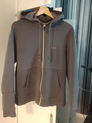 HUGO BOSS Grå huvtröja, Utmärkt skick. - Snygg grå hoodie från BOSS med dragkedja framtill och två stora fickor. Justerbar huva med snören, diskret BOSS-logga på bröstet Perfekt för en avslappnad och stilren look. Utmärkt skick och fräsch. 