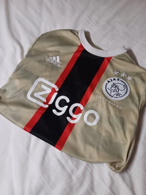 Ajax beige matchtröja Adidas M - Ajax matchtröja i beige med svart och röda ränder, tillverkad av Adidas. Tröjan har Ajax-logga, Ziggo sponsortryck och vita detaljer vid krage och axlar. Materialet är lätt och andas, perfekt för fotboll eller träning.