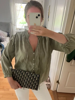 Olivgrön blus med knappar - Snygg olivgrön blus med v-ringning, knappar framtill och rynkad krage. Blusen har långa ärmar med resår vid ärmslut och ett avslappnat, luftigt fall. Perfekt att styla med jeans eller kjol för en trendig look.