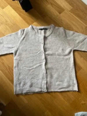 Ljusbeige basic kofta med knappar - En enkel och stilren ljusbeige kofta med rund halsringning och knäppning framtill. Koftan har långa ärmar och är tillverkad i ett mjukt, tunt stickat material som passar perfekt för lager på lager. Enkel att matcha till olika outfits.