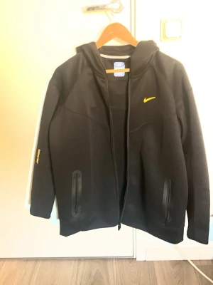 Svart Nike Nocta hoodie med gul logga - Säljer en svart hoodie från Nike med dragkedja och huva. Den har en gul Nike-logga på bröstet och diskreta fickor med dragkedja. Materialet känns mjukt och sportigt, perfekt för chill eller träning. Snygg detalj med gul text på ärmen.