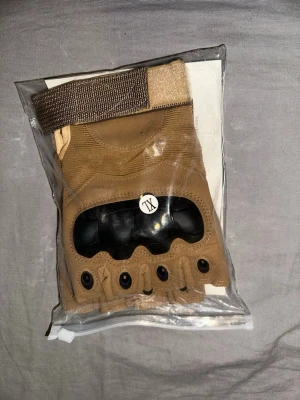 Beige fingerlösa träningshandskar - Säljer ett par beige fingerlösa handskar med förstärkt knogskydd i svart plast och justerbar kardborrestängning. Perfekta för träning eller utomhusaktiviteter där du vill ha extra grepp och skydd. Snygg och funktionell design. Storlek XL