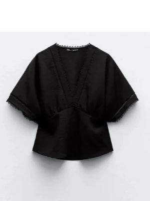 Svart blus med v-ringning och spetsdetaljer - Snygg svart blus med djup v-ringning och spetsdetaljer längs halsringning och ärmslut. Blusen har vida, korta ärmar och en avslappnad passform. Perfekt att matcha med jeans eller kjol för en trendig look.