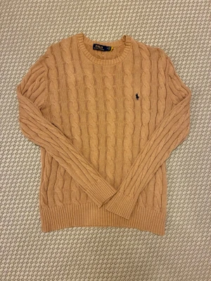 Beige kabelstickad tröja Polo Ralph Lauren - Kabelstickad tröja i beige från Polo Ralph Lauren med klassisk rund halsringning och broderad logga på bröstet. Tröjan har långa ärmar och ribbade muddar vid ärmslut och nederkant. Perfekt för en stilren och avslappnad look.