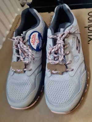 NYA Grå/rosa sneakers från Skechers stl 38 - Snygga grå/rosa sneakers från Skechers med mesh-ovandel och detaljer i ljusrosa och svart. Skorna har rosa- och svartmönstrade skosnören, extra mjuk innersula och en svartprickig sula med rosa inslag. Perfekta för en sportig och avslappnad stil. Vattentäta! Helt nya och oanvända. Se bilder!