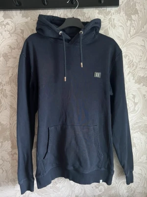 Mörkblå hoodie från Les Deux - Snygg mörkblå hoodie med klassisk känguruficka och justerbar huva med dragsko. Liten patch med 'II' på bröstet ger en clean look. Tillverkad i mjukt bomullstyg och har ribbade muddar vid ärmslut och nederkant. Skiftningarna i färg på bilden beror på ljuset genom kameran och är inget som syns i verkligheten. Hoodien är använd men är i fint skick!