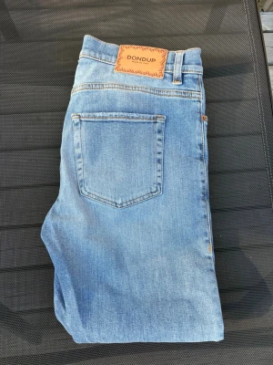 Ljusblå raka jeans från Dondup - Säljer ett par snygga ljusblå jeans från Dondup. femficksmodell i mjuk denim med läderpatch bak i midjan. Perfekta för en avslappnad och trendig look. Lite mindre slitningar som en del av designen.