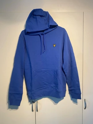 Lyle&Scott Hoodie blå - Snygg blå hoodie med huva från Lyle&Scott. Klassisk känguruficka framtill och ribbade muddar vid ärmar och nederkant. Perfekt för en chill och avslappnad stil. Storlek M och true to size. Knappt använd, mycket bra skick. 
