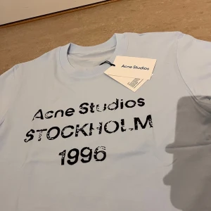 Ljusblå Acne Studios t-shirt - Snygg ljusblå t-shirt från Acne Studios med tryck i svart text framtill: 'Acne Studios STOCKHOLM 1996'. Klassisk rund hals och korta ärmar. Perfekt för dig som gillar stilrena plagg med en twist.
