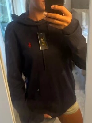 Mörkblå hoodie med röd logga - Snygg mörkblå hoodie från Polo Ralph Lauren med klassisk röd broderad logga på bröstet. Hoodien har huva med dragsko och långa ärmar. Perfekt för en avslappnad och trendig look. Materialet känns mjukt och bekvämt.lite tunnare i materialet än en vanlig hoodie!