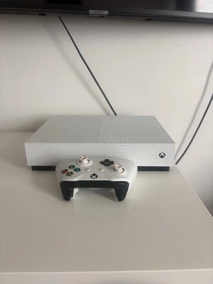 Vit Xbox One S med handkontroll - Säljer en stilren vit Xbox One S med matchande vit handkontroll. Konsolen har en rektangulär form med ventilerad ovansida och svarta detaljer. Perfekt för gaming och streaming, snygg design som passar in i alla rum.