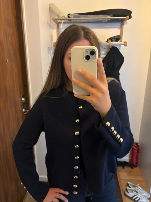 Mörkblå kavaj med guldknappar - Stilren mörkblå kavaj med raka axlar och dekorativa guldknappar längs hela framsidan och vid ärmsluten. Kavajen har en klassisk, rak passform och är utan krage för en modern look. Perfekt att styla till jeans eller kjol.