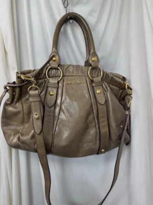 Miu Miu brun handväska i läder - Snygg handväska från Miu Miu i brunt läder med guldfärgade metalldetaljer. Väskan har både handtag och axelrem, samt dekorativa spännen och ringar. Perfekt för dig som gillar en lyxig och trendig look.