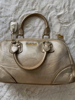 Guldig handväska från Miu Miu - Snygg guldig handväska från Miu Miu i äkta läder. Väskan har två handtag, guldfärgade metalldetaljer och ett dekorativt lås på sidan. Klassisk och stilren form som passar till många olika looks.