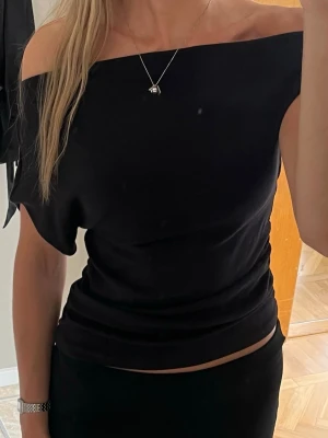 Svart offshoulder topp - Snygg svart offshoulder topp med tight passform och croppad längd. Toppen är ärmlös på ena sidan och har en stilren, enkel design som passar perfekt till många olika outfits. Tillverkad i mjukt material som sitter skönt mot kroppen.