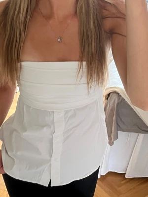 Zara topp - Säljer en vit axelbandslös tubtopp med knappar framtill. Toppen har bandeau-modell och är sydd i ett lätt bomullstyg. Perfekt för dig som vill ha en clean och stilren look med en twist.