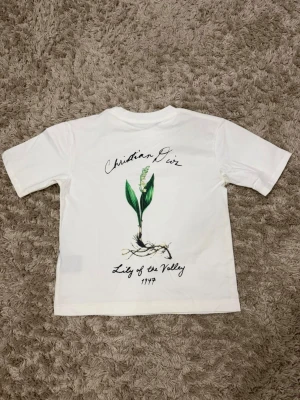 Vit Christian Dior t-shirt med tryck - Vit t-shirt från Christian Dior med ett stilrent tryck av liljekonvalj och texten 'Lily of the Valley 1947' på ryggen. Framtill finns en liten diskret logga på bröstet. Klassisk passform och rund hals. Perfekt för dig som gillar exklusiva och unika plagg.