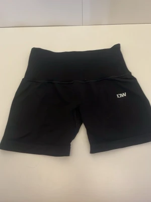 Svarta högmidjade träningsshorts - Snygga svarta högmidjade shorts med DW-logga på vänster ben. De är tillverkade i ett stretchigt syntetmaterial som passar perfekt för träning. Modellen har en bred linning för extra komfort och stöd.