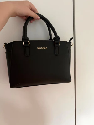 Svart handväska från Don Donna - Stilren svart handväska från Don Donna med två handtag och guldfärgade detaljer. Väskan har en klassisk, rektangulär form och är tillverkad i slätt konstläder. Perfekt för dig som gillar enkel och tidlös design.