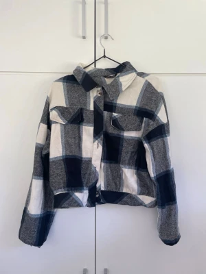 Blåvitrutig overshirt med fickor - Snygg overshirt i blått och vitt rutmönster. Modellen har två bröstfickor med lock, klassisk krage och knäppning framtill. Overshirten är tillverkad i ett mjukt tyg och har avslappnad passform med långa ärmar.
